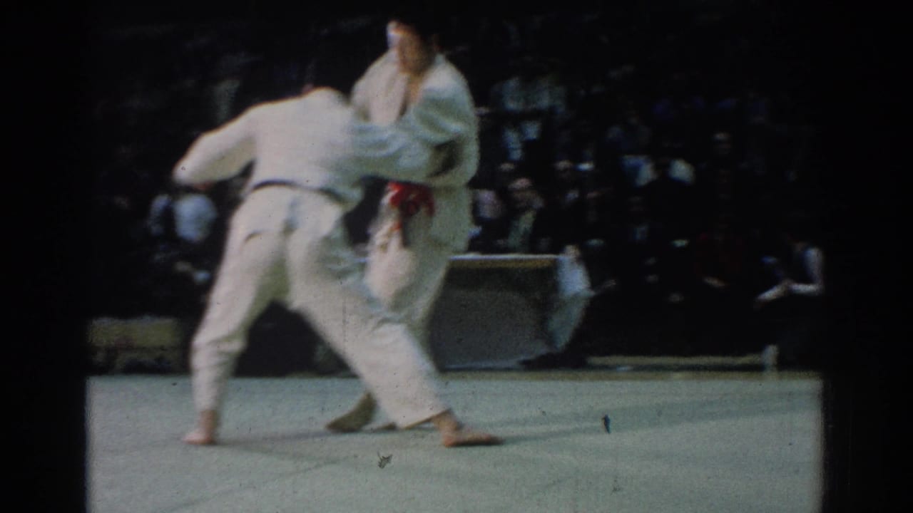 Two judo competitors in white gi grapple on a mat before a seated audience.