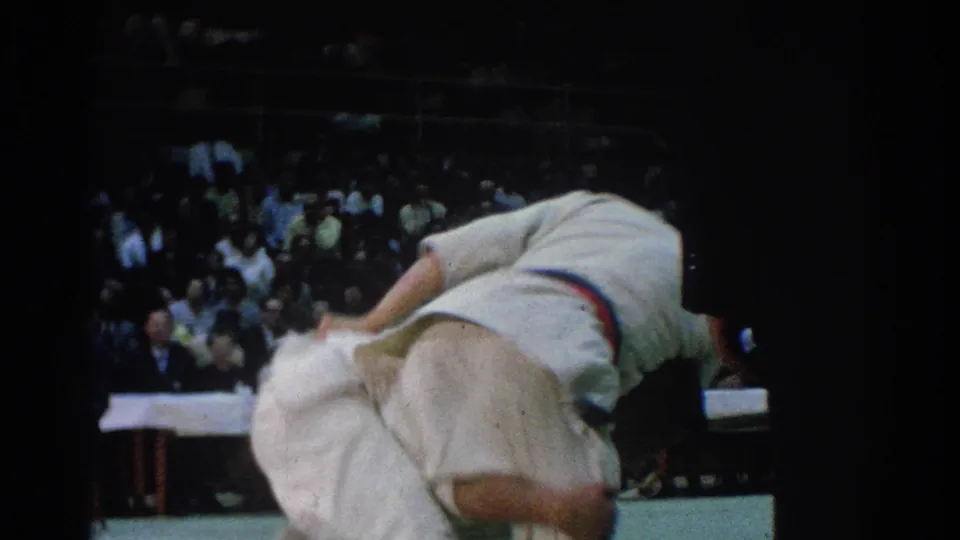 A competitor in a white gi with a red belt is bent low during a throw or takedown as spectators and officials sit behind the mat.