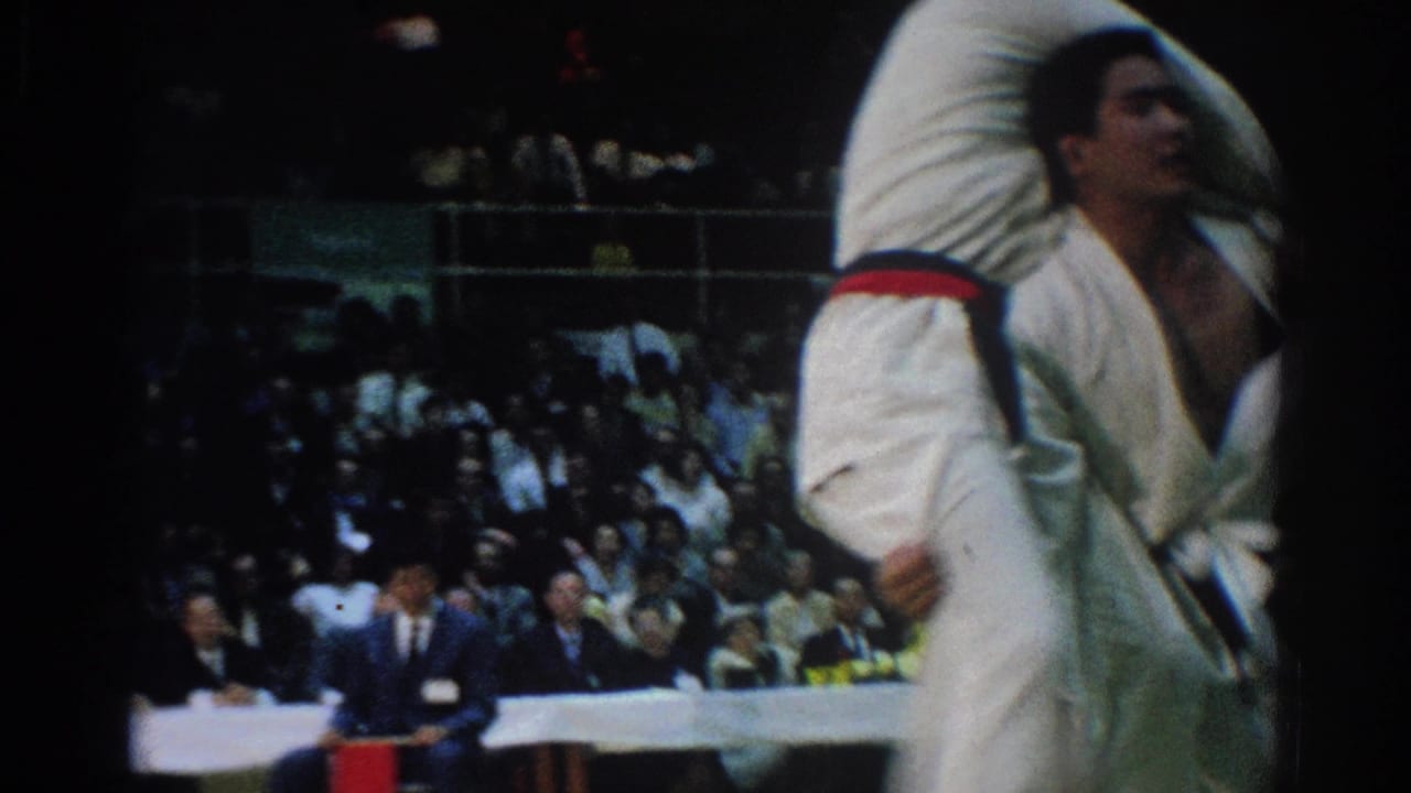 Judo competitor in white gi lifts another competitor during a throw before a crowded indoor audience.