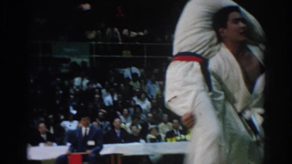 In West Covina, 1966, a male judo competitor in a white gi raises his opponent during a throw as spectators and judges watch.