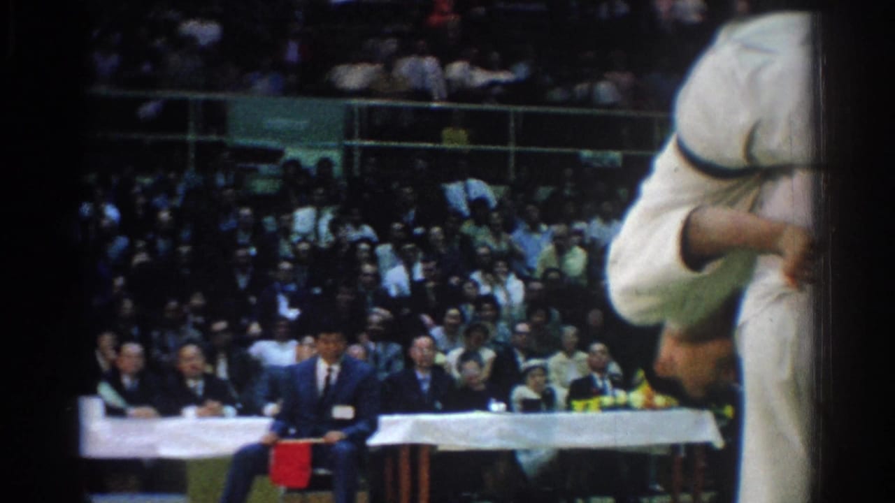 During the 1966 West Covina judo match, a competitor is thrown near the frame edge as officials sit at a long table behind the mat.