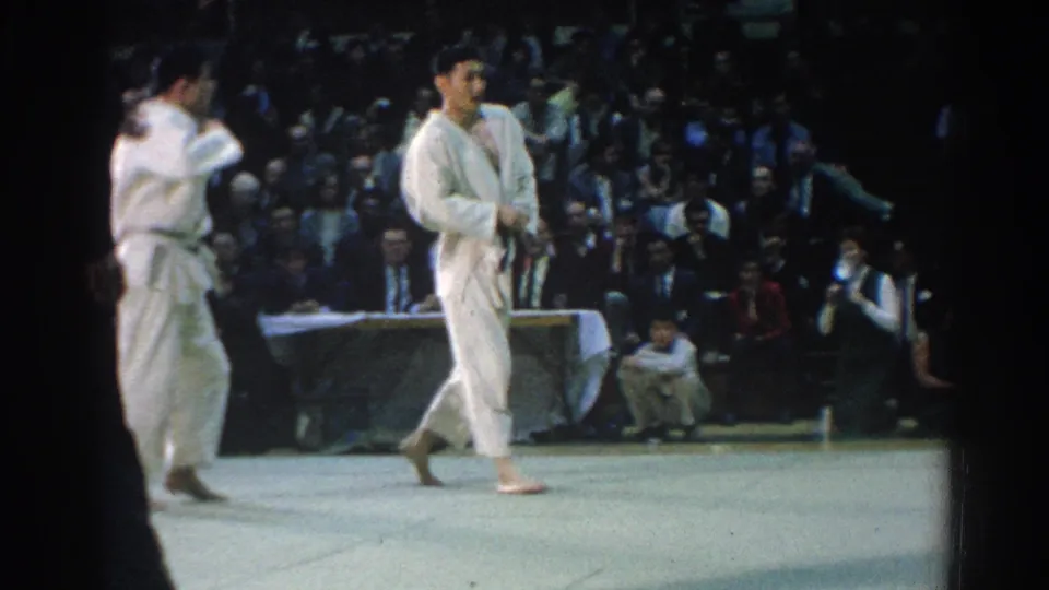 Two male judo competitors reset on the mat in a crowded West Covina indoor arena, with the 1960s home-movie camera shooting from audience level.