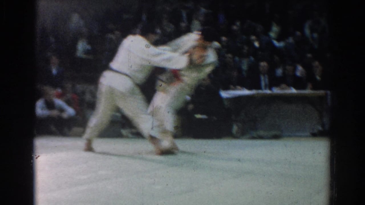 Two men in white judo gis grapple during a 1966 indoor match in West Covina, with spectators behind the mat.