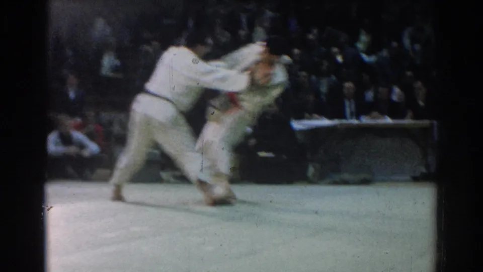 Two men in white judo gis grapple during a 1966 indoor match in West Covina, with spectators behind the mat.