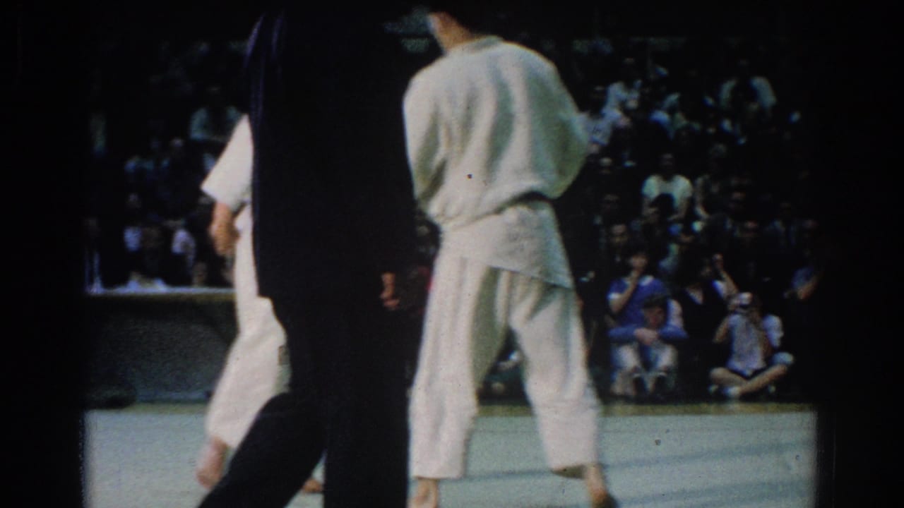 During the West Covina judo match, one competitor stands on the mat while an official and crowd are visible in the background.