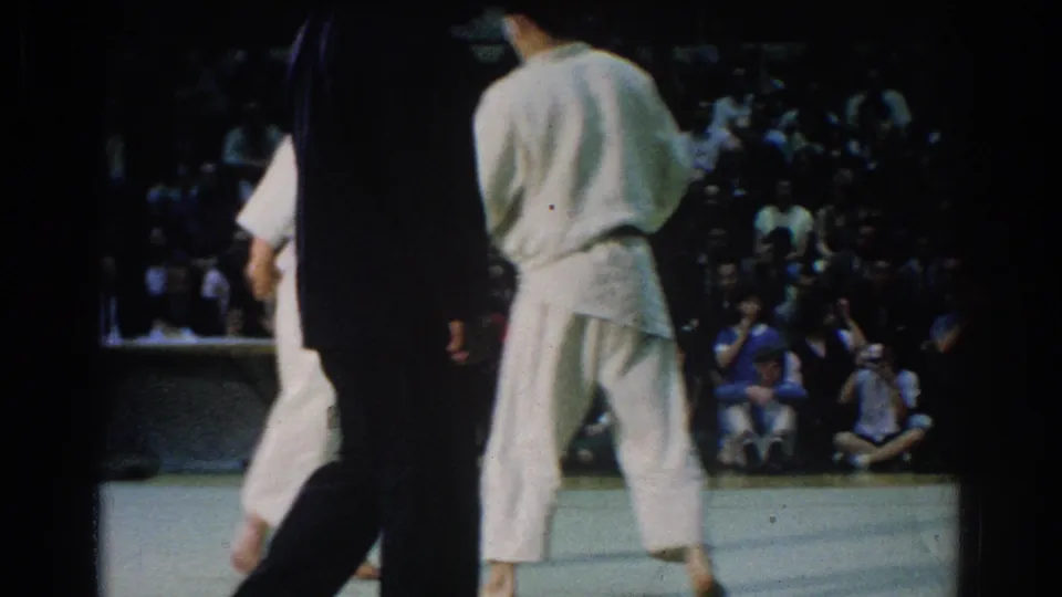 During the West Covina judo match, one competitor stands on the mat while an official and crowd are visible in the background.