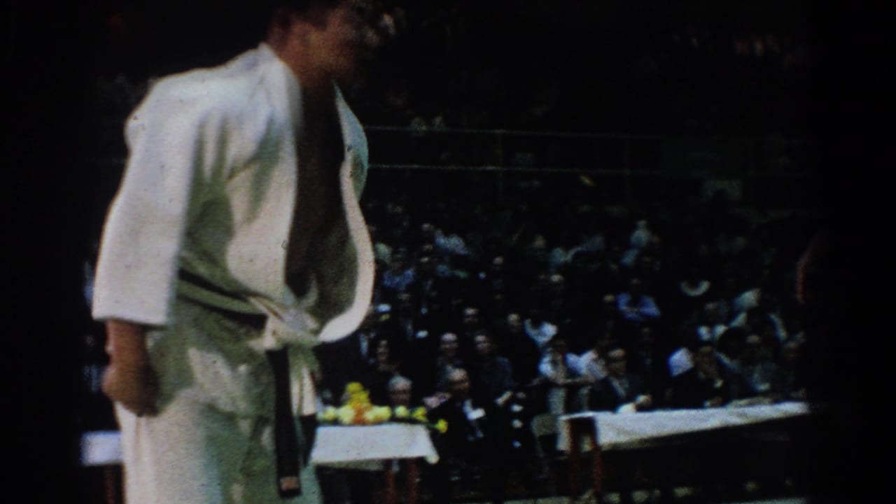 Referee or official in white uniform stands near a judo mat with spectators in dark bleachers behind him.