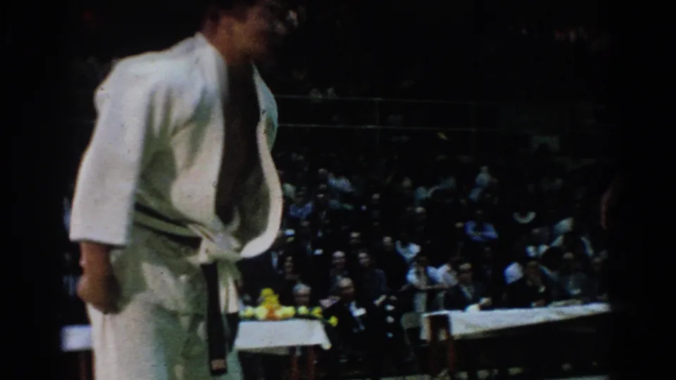 In the 1966 West Covina judo tournament footage, an official in a white uniform stands near the mat while a packed indoor audience watches from shadowed seating.