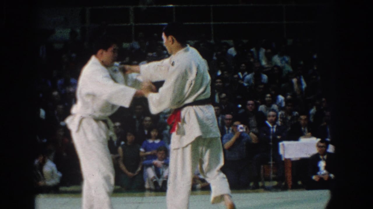 Two judo competitors in white gi grip each other on the mat in front of a seated crowd.