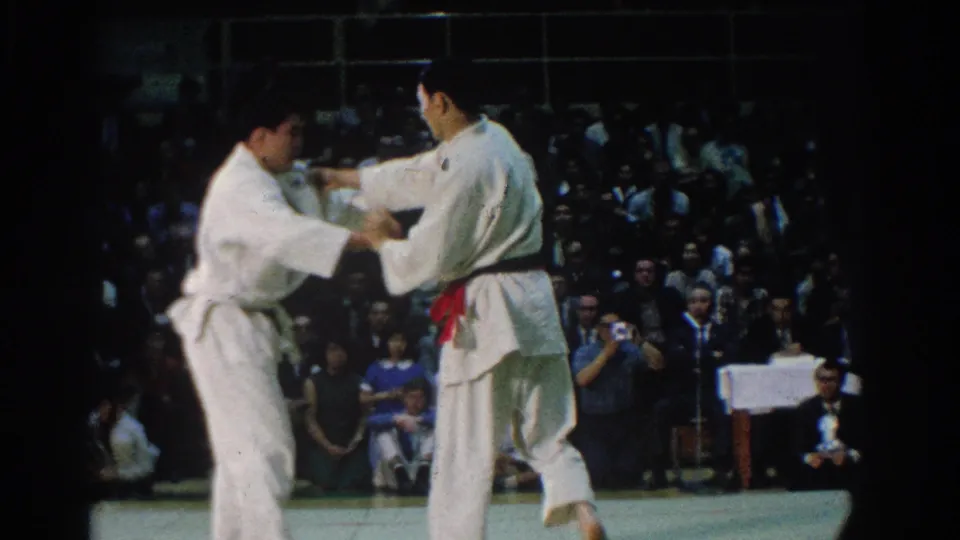 Two competitors in white gi, one wearing a red belt, grip each other during a judo match inside the West Covina arena setting described by the clip metadata.