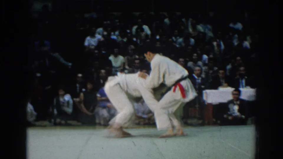 A judo competitor in a red belt works from a standing grip as the opponent bends low, with the crowd visible behind the mat in the 1966 tournament footage.