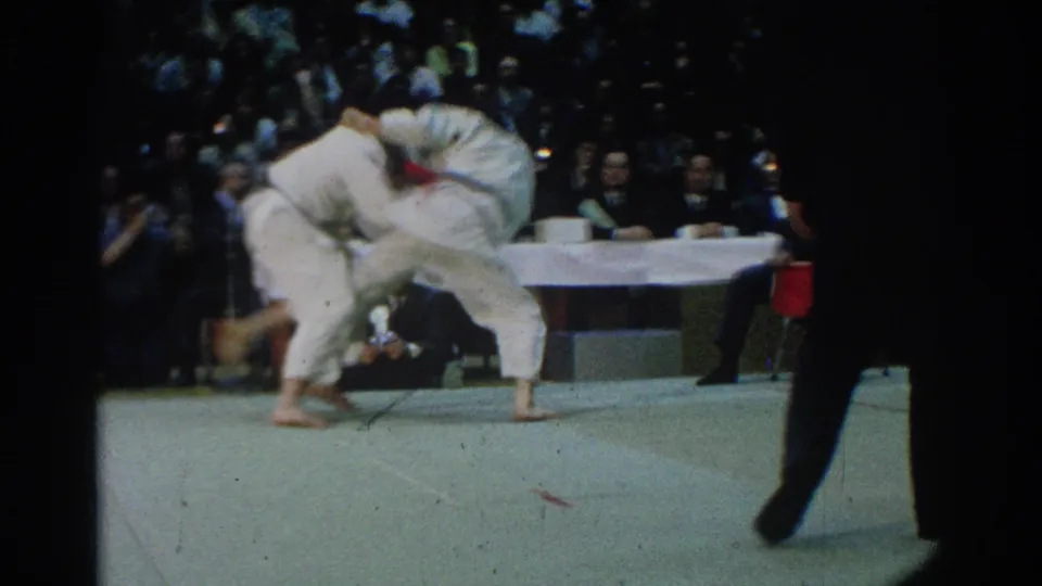 The match reaches a dynamic throw moment, with one competitor turning the other across the mat while an official observes from the side and spectators watch behind them.