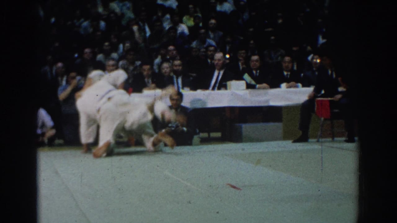 Two judo competitors in white gi tumble on a mat as judges and spectators watch behind them.