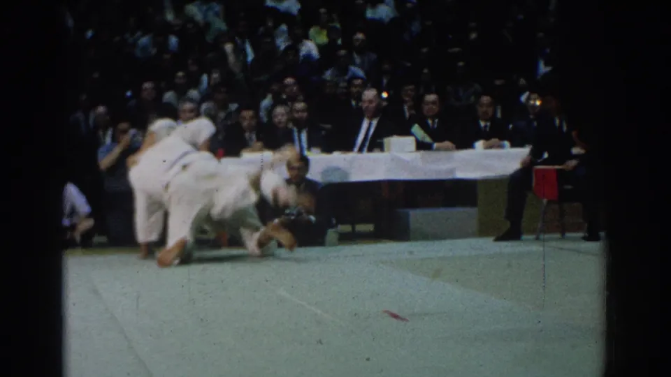 In West Covina, California in 1966, two judo competitors in white gi fall across the mat during a tournament before seated judges and a crowd.