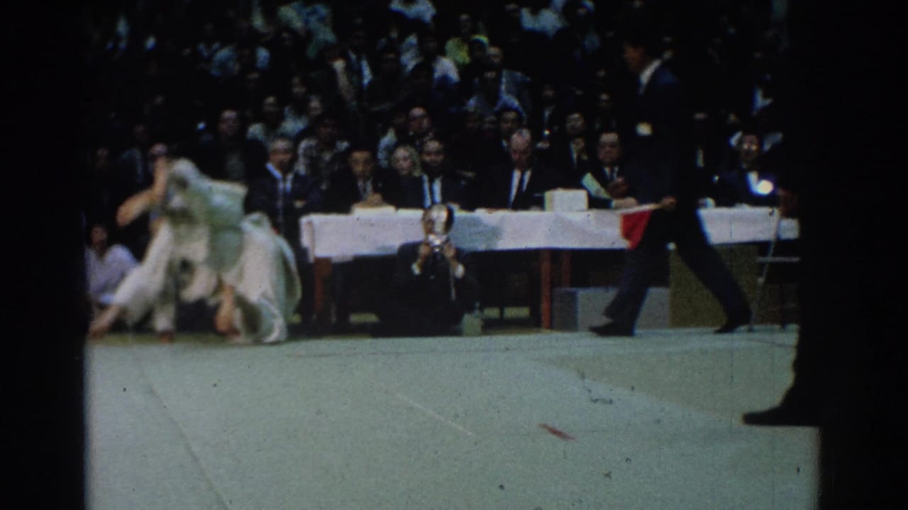 A 1966 West Covina judo match continues as one competitor rises from a throw, with judges seated behind and a referee crossing the edge of the frame.