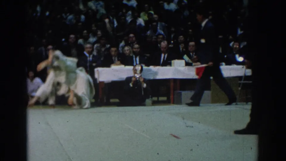 A 1966 West Covina judo match continues as one competitor rises from a throw, with judges seated behind and a referee crossing the edge of the frame.