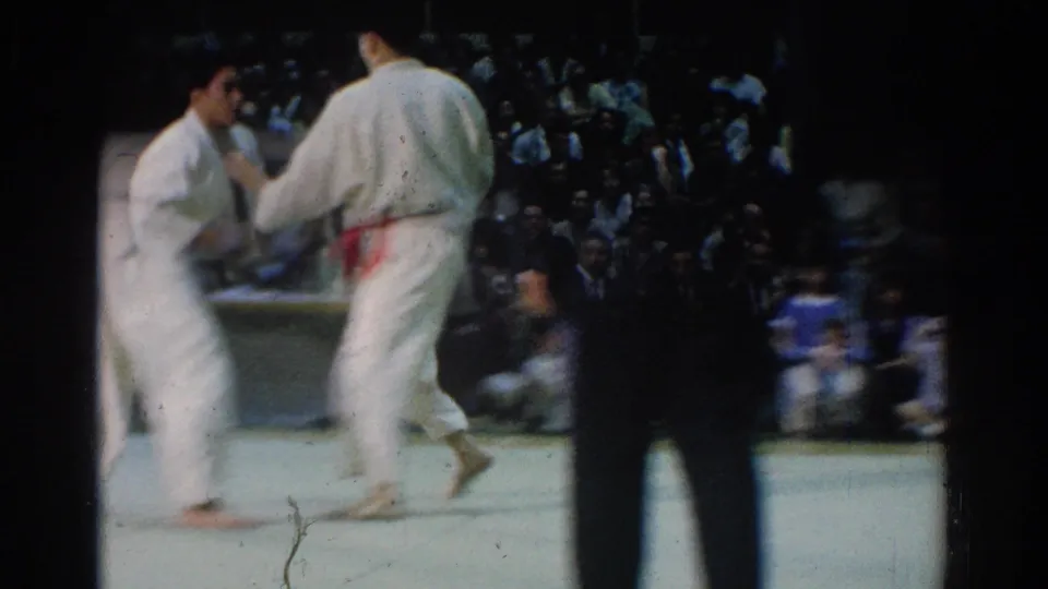 During the West Covina tournament, the two competitors stand close on the mat in white gi while a crowd watches from the dim background.