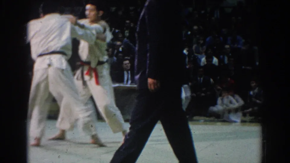 Two judo competitors in white gi engage on the mat in 1966 West Covina as a dark-clothed referee passes in front of the camera.