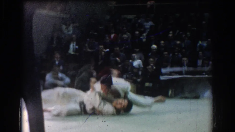 1966 West Covina 8mm frame showing a judo throw in progress on an indoor mat with spectators seated behind the action.