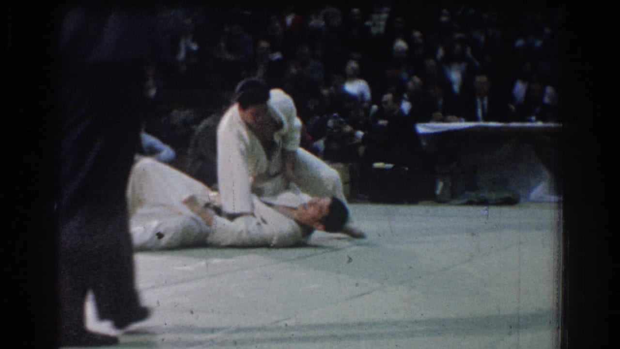 1966 West Covina judo match frame with one athlete above another on the mat while an official or passerby is partly visible nearby.