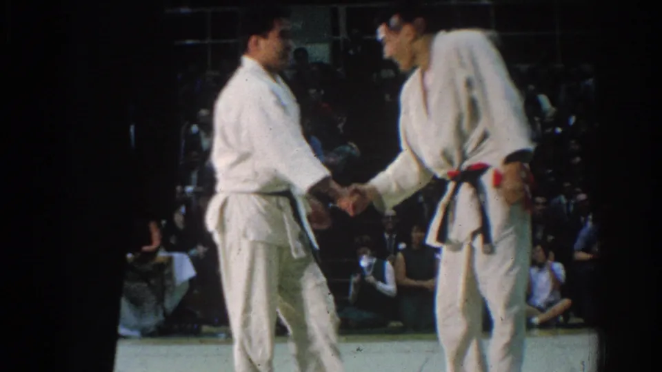 1966 West Covina 8mm frame showing the two judo athletes shaking hands on the mat with spectators in the background.
