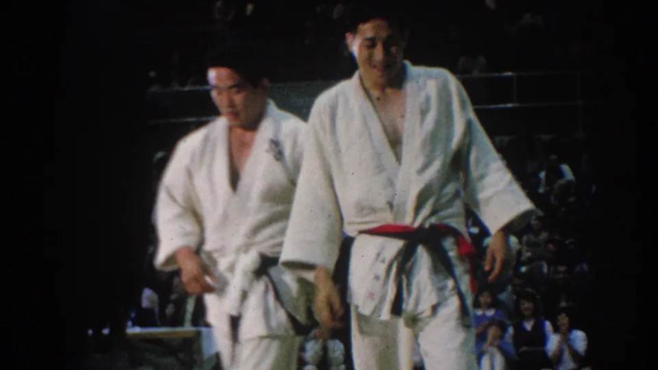 1966 West Covina frame showing two judo competitors leaving the mat, one wearing a red belt, with the audience behind them.