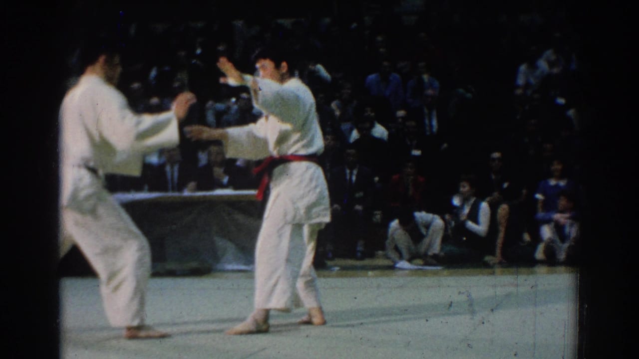 Two karate competitors in white gi face each other on a mat before seated spectators.