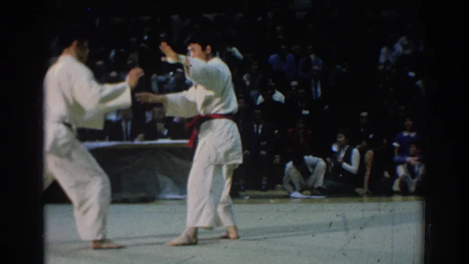 In 1966 West Covina footage, two karate competitors face off on a mat while a seated crowd watches from the background.
