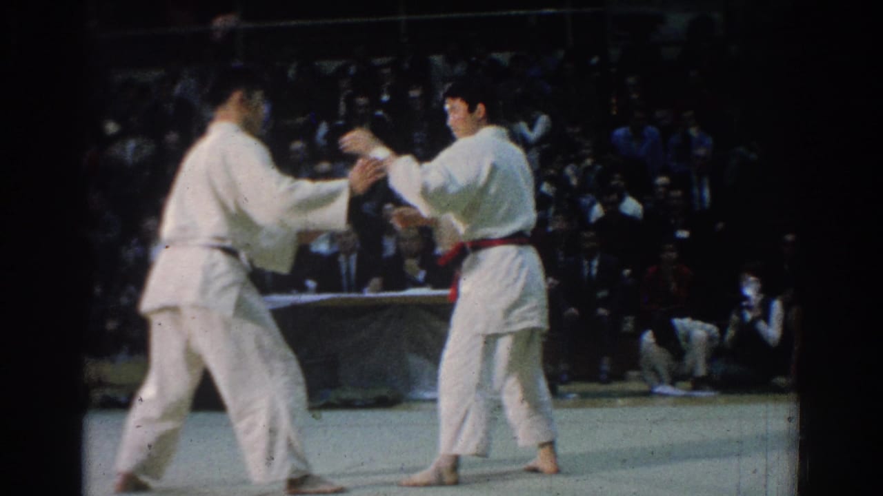 Two white-gi competitors move into contact during a 1966 West Covina karate match, with the audience dimly visible behind them.