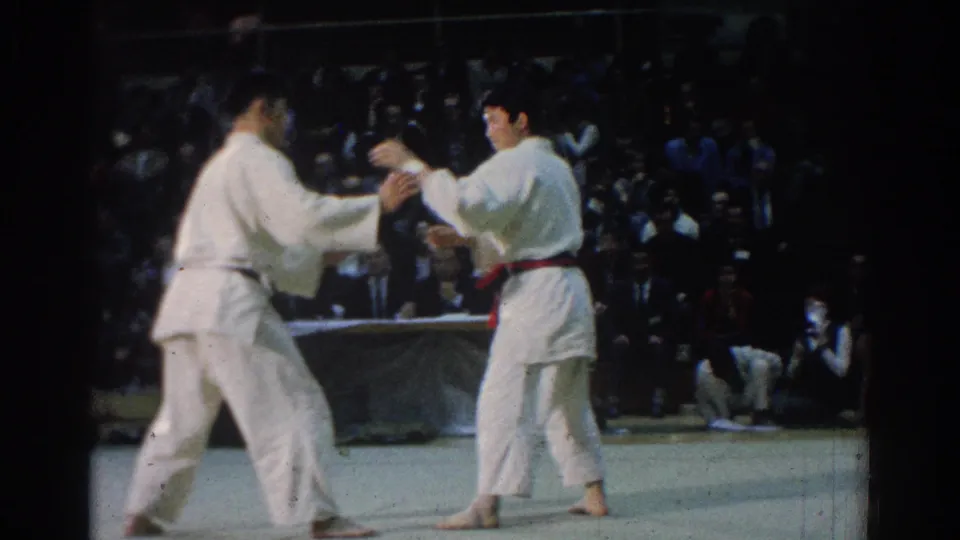 Two white-gi competitors move into contact during a 1966 West Covina karate match, with the audience dimly visible behind them.