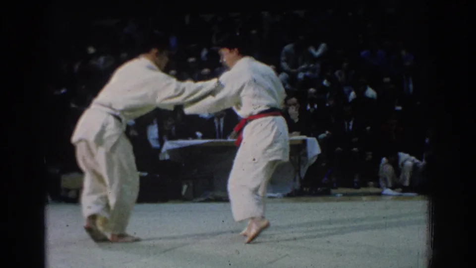 During the West Covina match, the competitors lean forward in a close karate exchange while spectators sit along the far side of the mat.