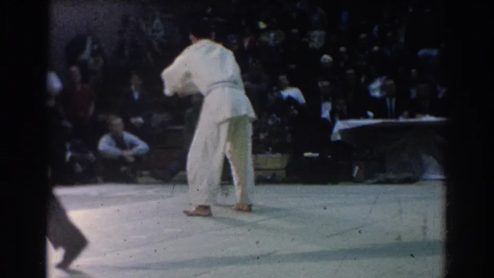 A competitor in a white gi stands turned away near the mat edge in the 1966 West Covina karate footage, with the crowd in soft focus behind.