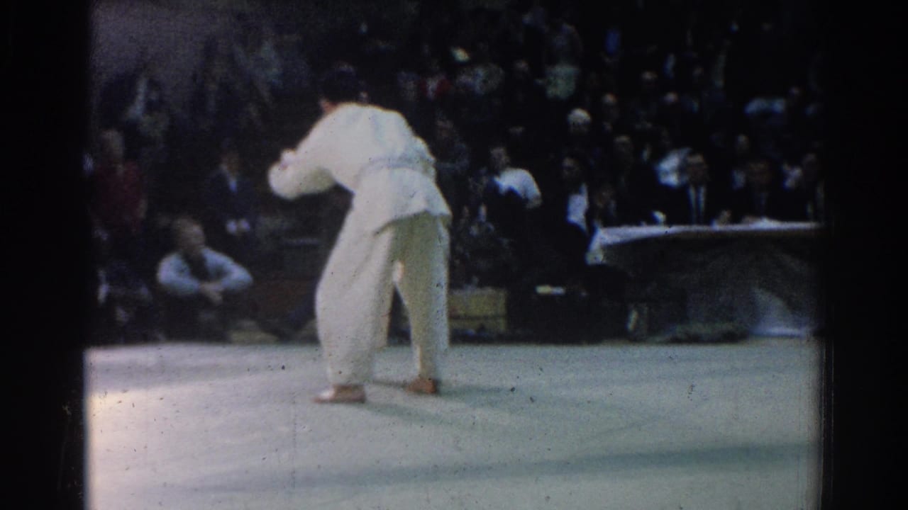 A martial arts competitor in a white gi stands on a mat before a seated audience.