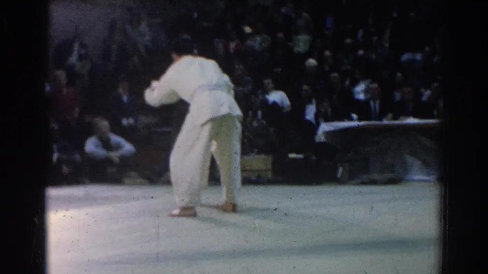 A gi-clad competitor appears on a mat during a 1966 West Covina martial arts match, with spectators and an officials' table in the background.