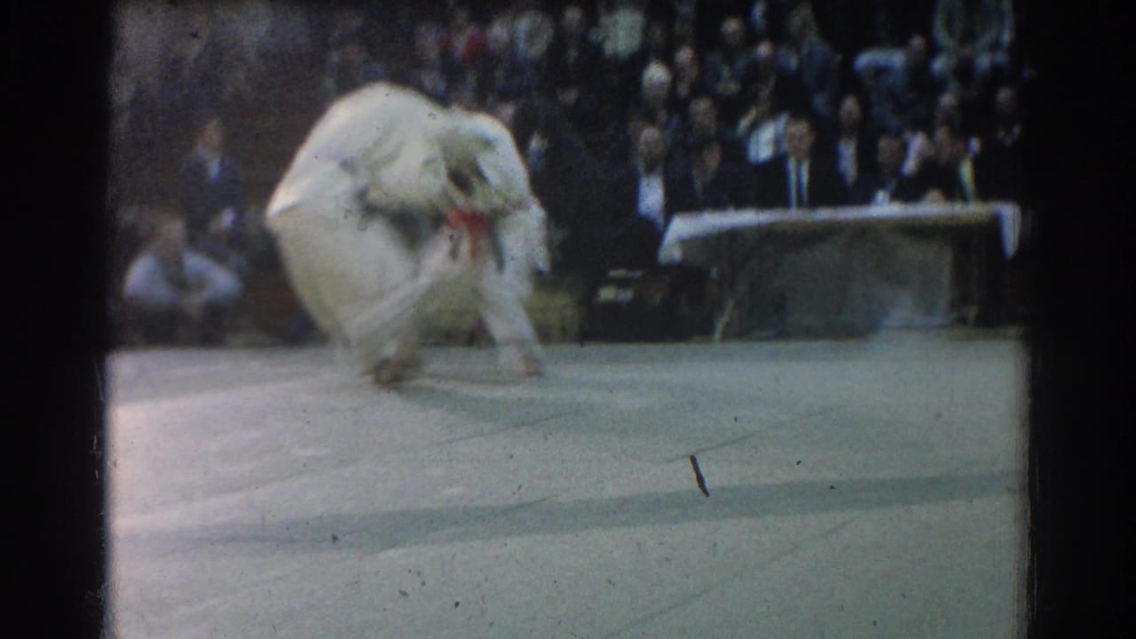 During the West Covina match, a competitor in a white gi drops into a low motion on the mat while spectators sit behind the action.