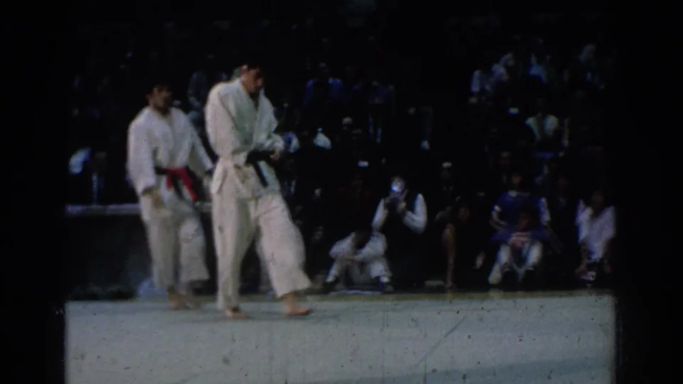 Two competitors in white gis stand together on the mat at the West Covina event, with audience members and officials visible behind them.