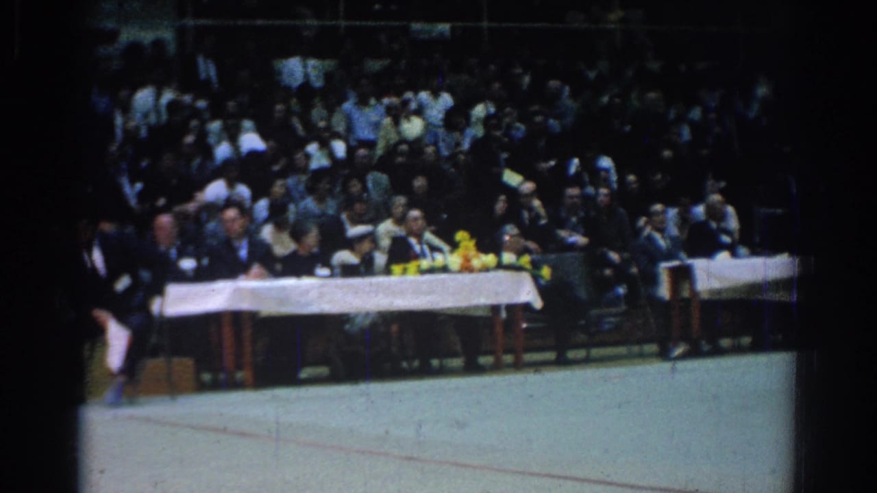 Judges sit behind a long white-covered table as a crowd watches from gym bleachers.