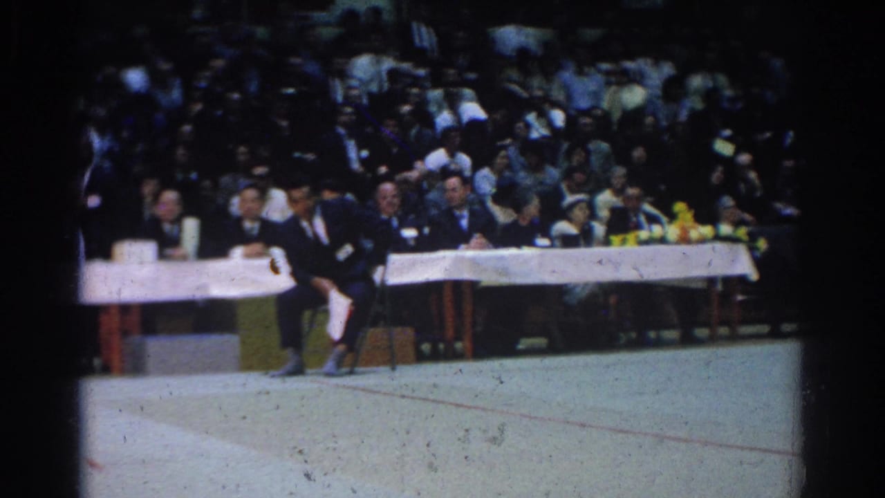 A competitor in dark clothing stands near the judges table before a seated audience.