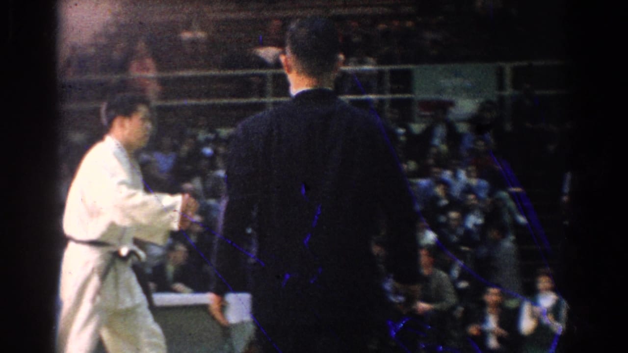 1966 West Covina judo match frame showing a white-gi competitor near a dark-suited referee before a crowded gym audience.