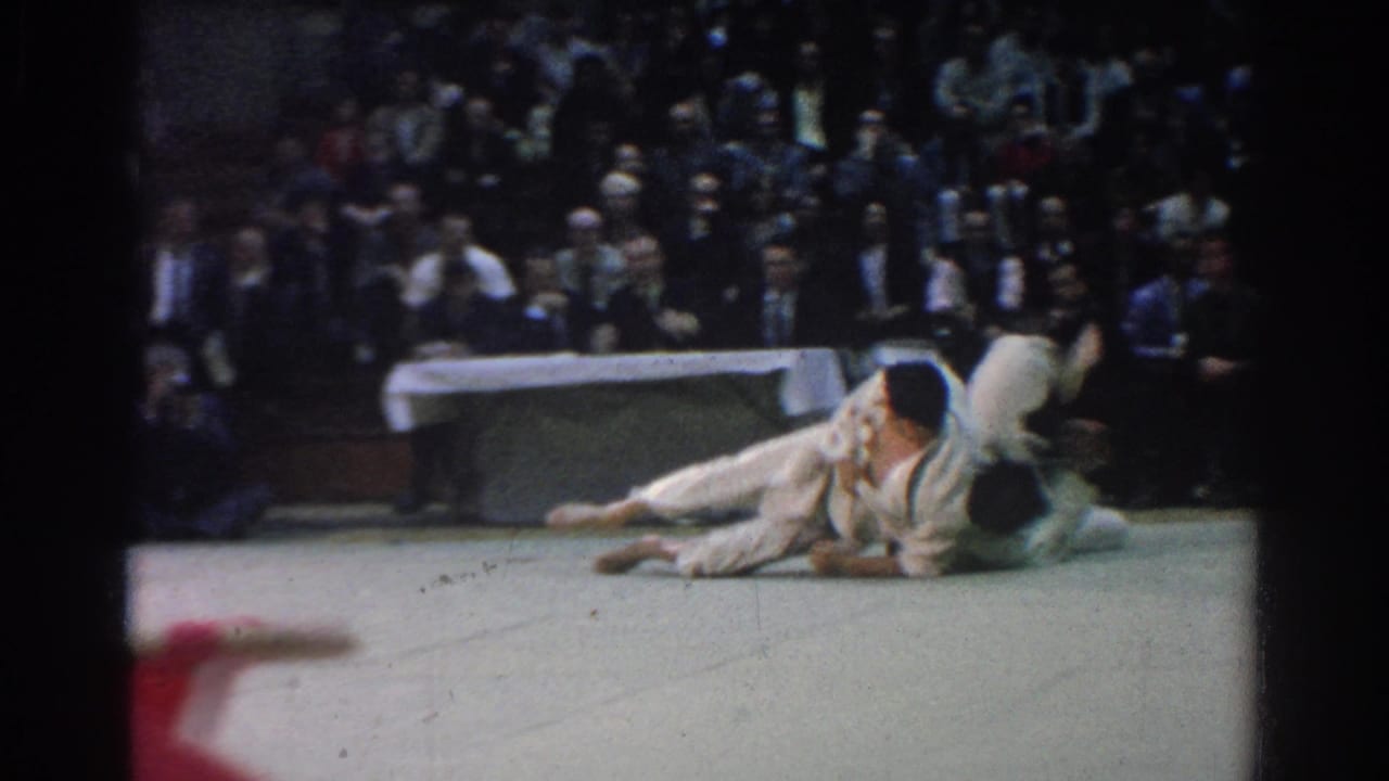 1966 West Covina judo match frame showing two competitors on the mat during a ground exchange before seated spectators.