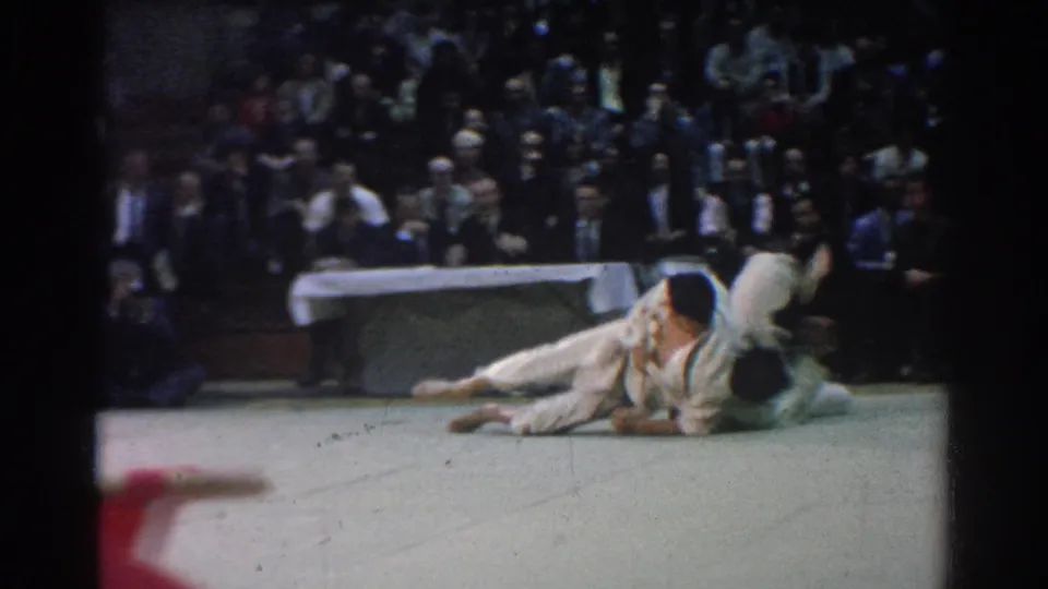 1966 West Covina judo match frame showing two competitors on the mat during a ground exchange before seated spectators.