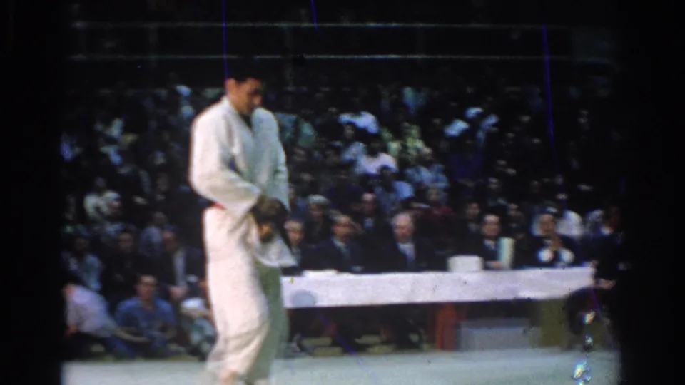1966 West Covina judo match frame showing a white-gi competitor standing after an exchange, with the gym audience behind him.
