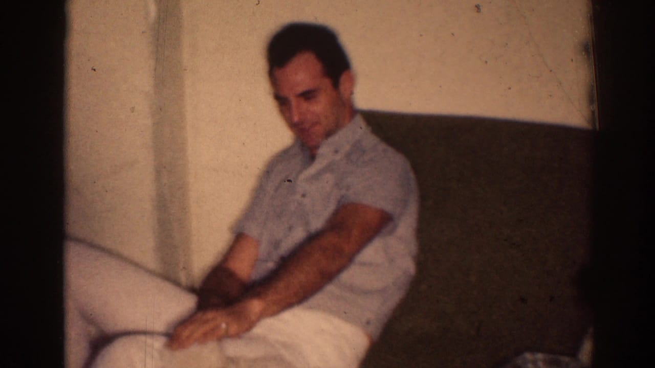 Adult man in a light shirt seated on a sofa against a plain wall.