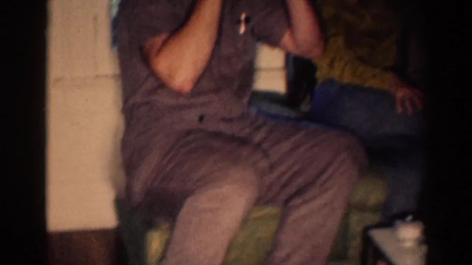 In the 1967 Los Angeles home movie, a seated man brings sunglasses up while another person is partly visible nearby.