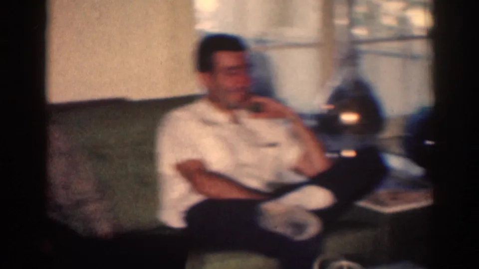 Adult man reclines on a sofa in the 1967 Los Angeles interior, with a lamp and tabletop objects beside him.