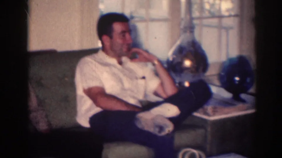 1967 Los Angeles home-movie frame of a man seated on a green couch, resting his chin on his hand near windows and decorative objects.