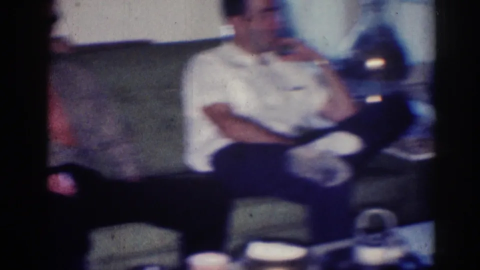 Soft 8mm frame from the Los Angeles interior showing a seated man on a couch, with low table items and living-room furnishings nearby.