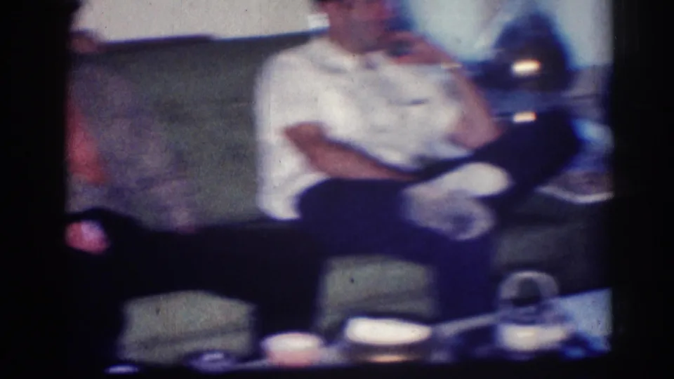 Soft, color-shifted 8mm view of the couch area, with a seated man, dark furniture, and cups visible in the private interior.