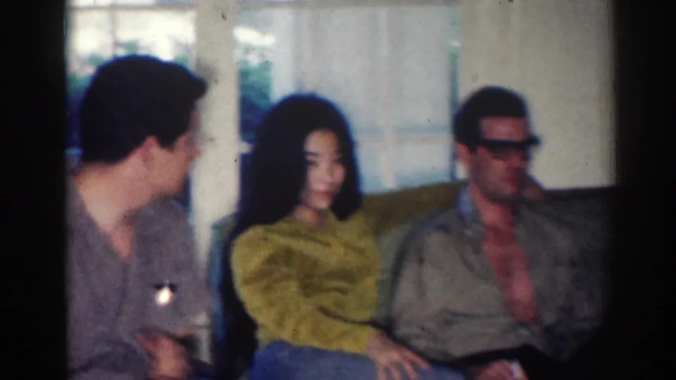1967 Los Angeles home-movie frame of three adults seated on a green couch, with a woman in a yellow sweater between two men.