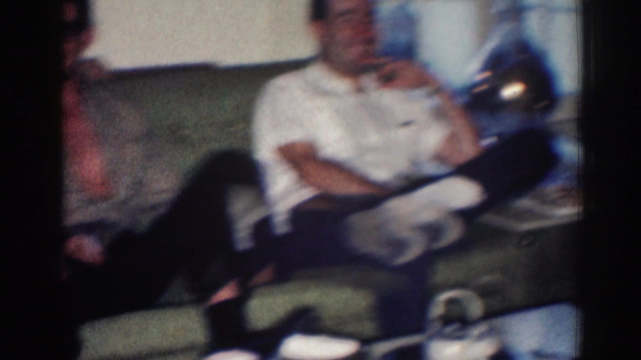 In this 1967 Los Angeles home-movie frame, a man in a white shirt sits on a couch in a dim living-room interior.